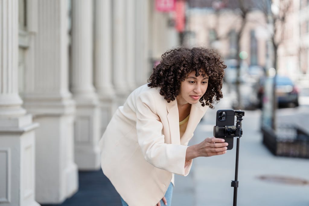 Attentive young ethnic female vlogger leaning forward to cellphone on tripod while preparing for video record in city