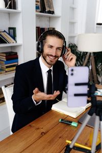 A confident businessman in a suit engaging in a video call from a home office featuring bookshelves and a smartphone tripod.