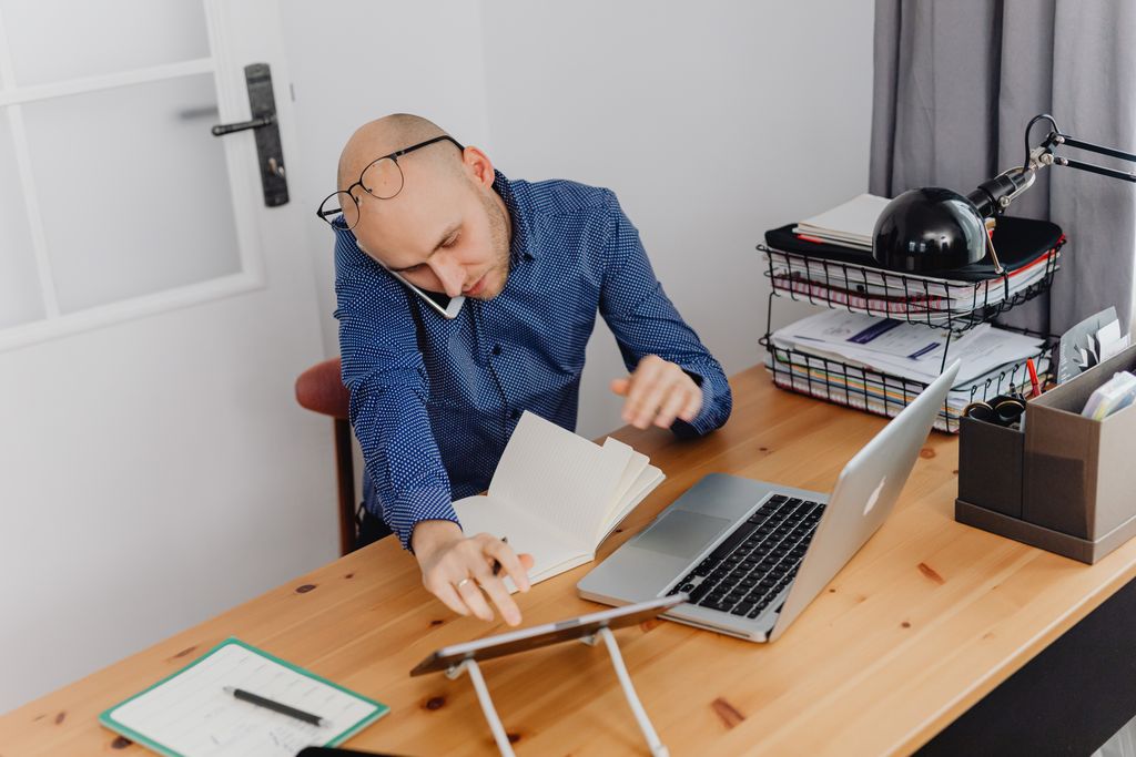 A man multitasking with phone, laptop, and notebook on his desk at home office.