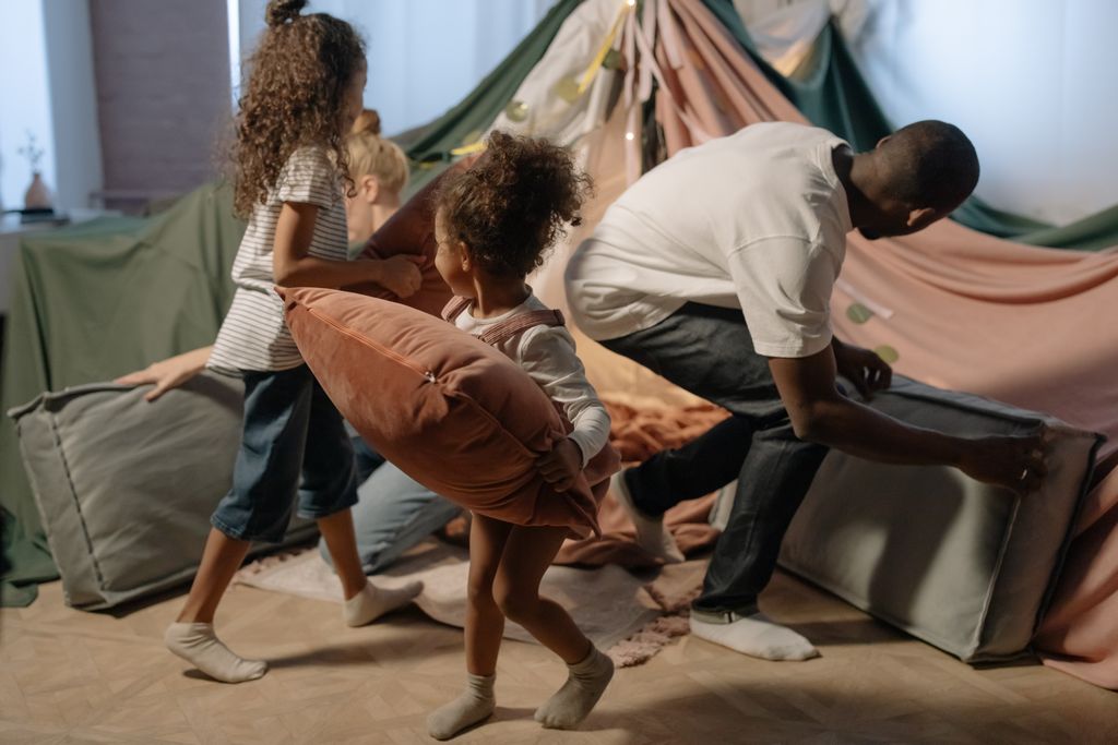 A playful family scene with a father and daughters having a pillow fight inside a cozy indoor fort.